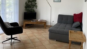 Ein Wohnzimmer mit grauem Sofa, einem schwarzen Sessel, einem Holztisch, einem Fernseher und einer Pflanze.