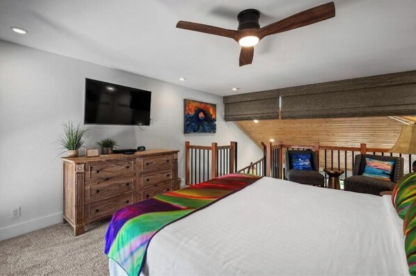 A bedroom with a bed, wooden dresser, TV, artwork, and a ceiling fan.