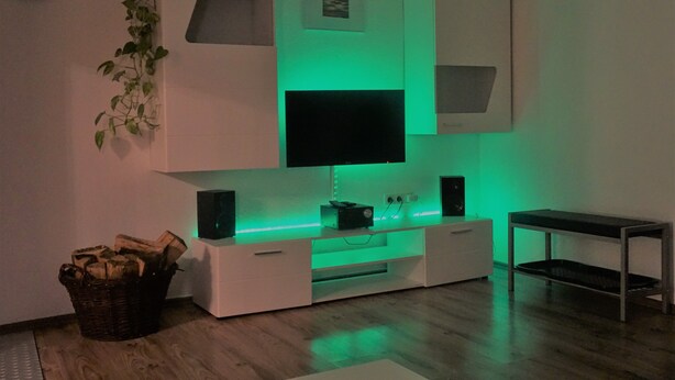 Un salon moderne avec un téléviseur à écran plat fixé à un meuble de télévision blanc, un éclairage d’ambiance vert, un plancher en bois, une petite table et une horloge murale.