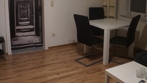 Une pièce avec un plancher en bois, une table blanche avec deux chaises, une petite table blanche avec des tasses, un ventilateur de plafond et une porte donnant sur un couloir.