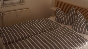 Une chambre à coucher avec un lit, une literie à rayures, une fenêtre avec des stores, un radiateur et une horloge fixée au mur.