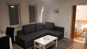 Un salon moderne avec un canapé d’angle gris, une table basse blanche et une petite table d’appoint blanche. On y trouve une fenêtre avec des stores, une porte donnant sur une chambre et un tapis gris sur le plancher en bois.