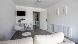 Ein modernes Wohnzimmer mit einer Couch, einem Esstisch mit Stühlen, einem an der Wand montierten Fernseher und einer Küche im Hintergrund.