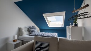 Ein Schlafzimmer mit schrägen Wänden, einem Oberlicht, einem Bett mit gestreifter Bettwäsche, einer weißen Kommode und einer hängenden Pflanze.