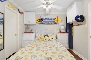 A bedroom with a bed, two nightstands, a ceiling fan, a surfboard, and a blue neck pillow.