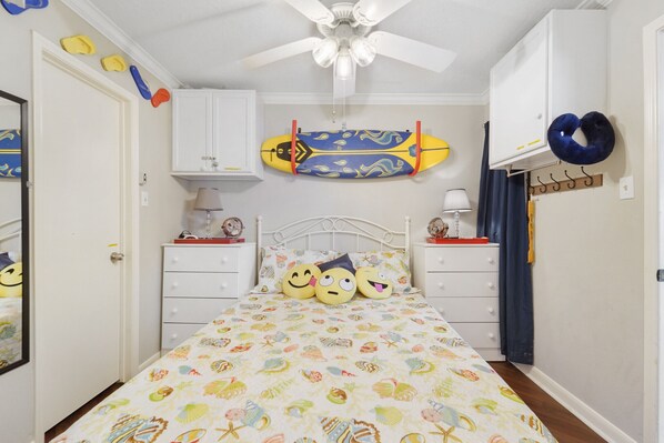 Une chambre avec un lit, deux tables de chevet, un ventilateur de plafond, une planche de surf et un coussin de voyage bleu.
