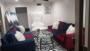 Ein Wohnzimmer mit einem weißen, gesteppten Sessel, zwei blauen Sofas mit roten Kissen und einem Glastisch sowie einem Zebra-Muster Teppich.