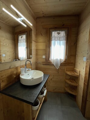 A wooden cabin bathroom with a round sink, a mirror, a window with curtains, and built-in shelves.