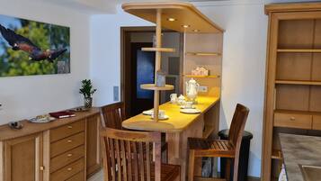 A dining area with a wooden table, chairs, a hanging light fixture, a wall-mounted painting of a bird, and a potted plant.