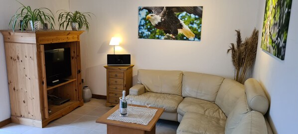 A living room with a beige sofa, wooden entertainment unit, and a TV. There are plants on a shelf, a lamp, and artwork on the wall.