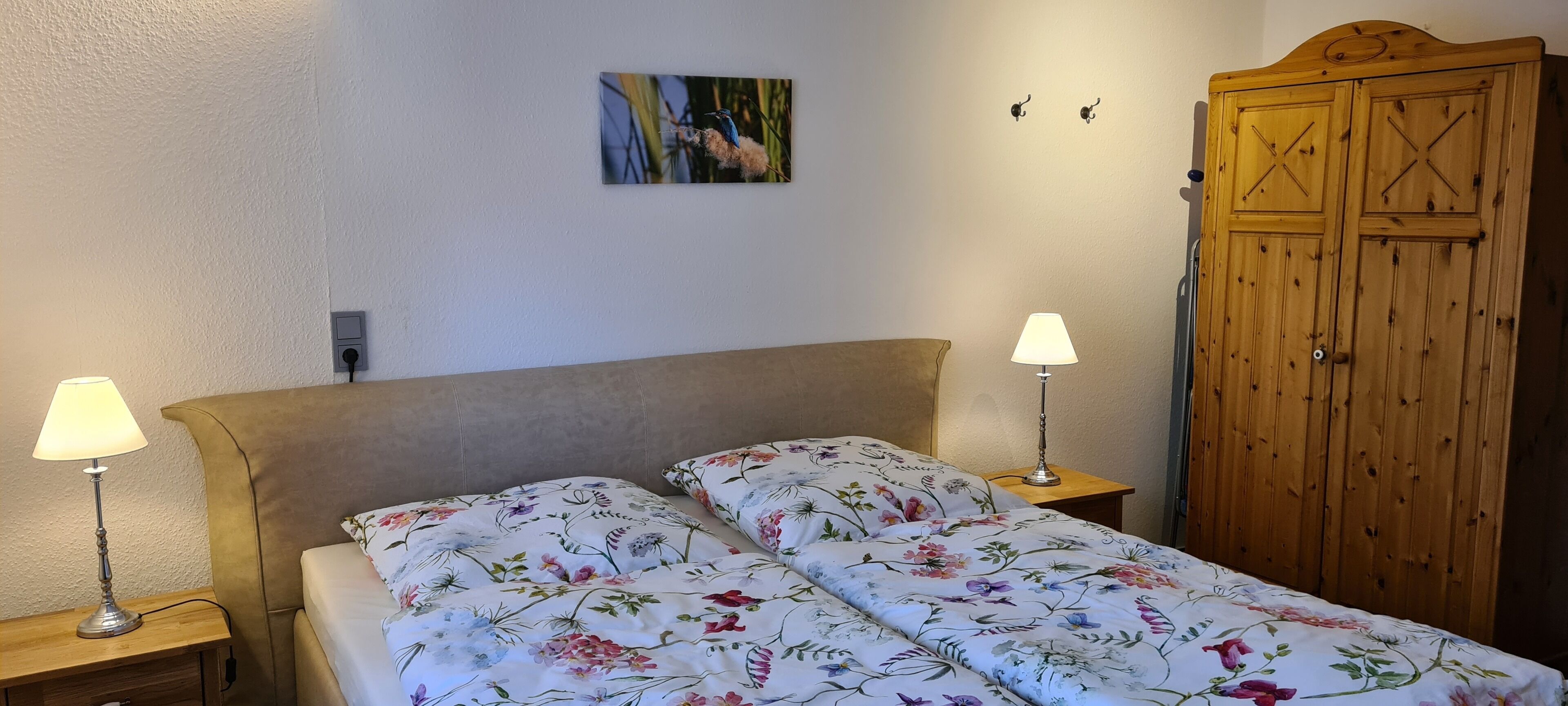 A bedroom with a bed, two bedside lamps, a wooden wardrobe, and a wall-mounted picture.