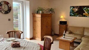 A living room with a beige sofa, wooden cabinet, dining table with chairs, and a wall clock. - 5-star vacation apartment "Hedfan" in Cnauceltaidd (Halle)
