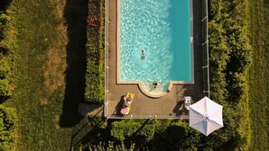 Vista aérea de una piscina rectangular rodeada por un muelle de madera, con una persona nadando y otra relajándose en una tumbona.