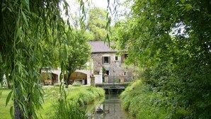 Una casa de piedra con un puente sobre un estrecho canal, rodeada de abundante vegetación.
