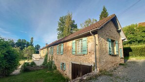 Una casa de piedra con techo inclinado y contraventanas verdes, rodeada de vegetación y un camino de grava.