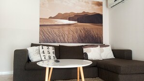 Un salon avec un canapé marron foncé, une table basse blanche aux pieds en bois et un grand tableau représentant un paysage de plage accroché au mur.