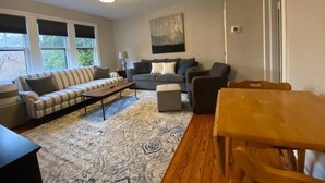 A living room with a sofa, armchairs, a coffee table, and a rug. There are windows with blinds, a painting on the wall, and a door. - Spectacular Location: State Circle Apartment (Annapolis)