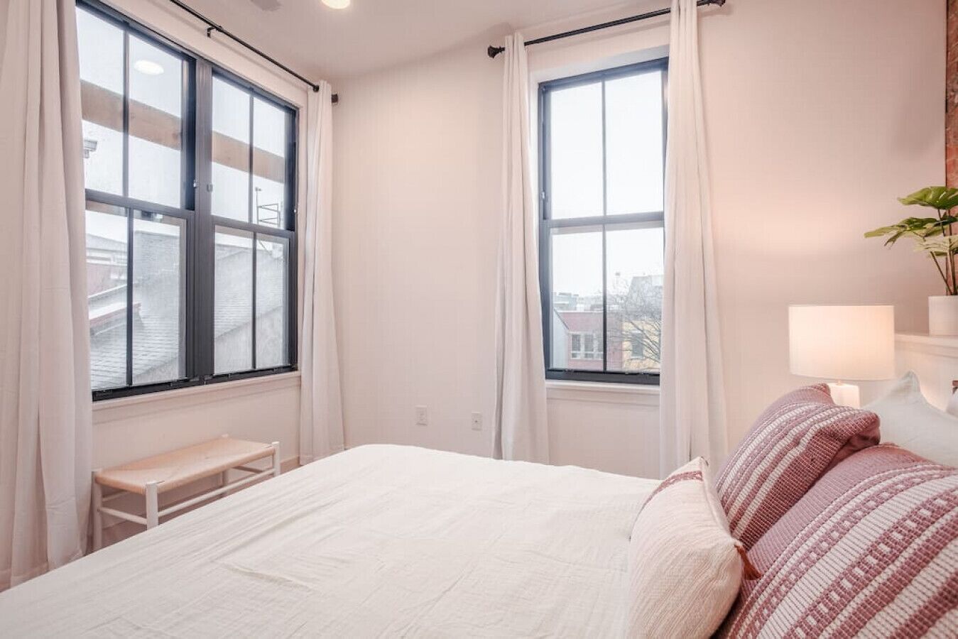 A bedroom with a bed, a bench, two windows with curtains, a lamp, and a plant.