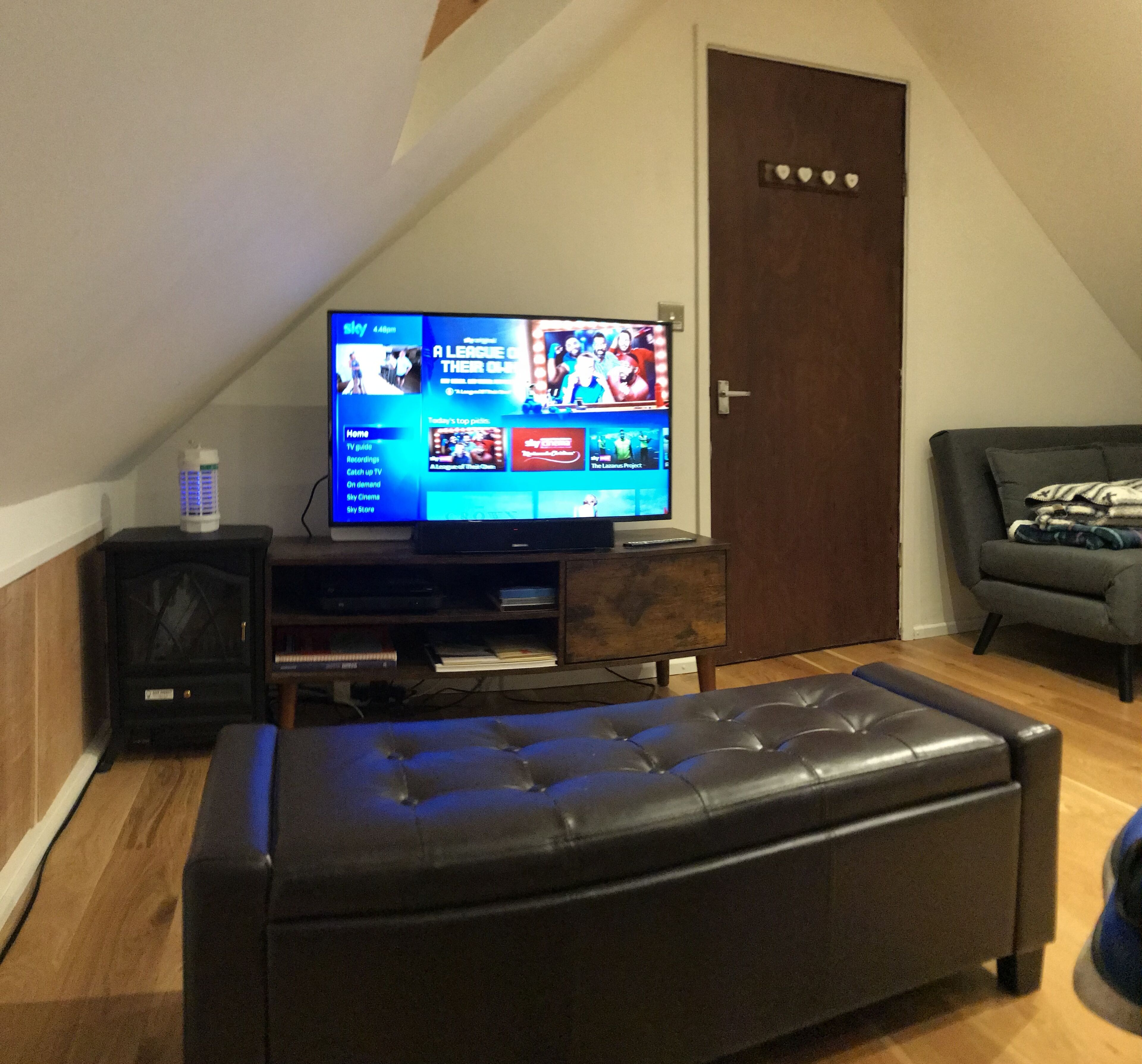 A living room with a flat-screen TV on a wooden stand, a black leather ottoman, a wooden cabinet, and a grey sofa.