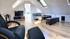 A modern loft apartment with a high ceiling, wooden flooring, a grey sofa, a black ottoman, a TV on a wooden stand, and skylights. - Cosy new Loft with bathroom and facilities (Glynneath)