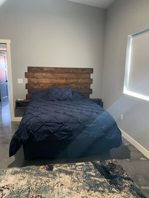 Un dormitorio con cabecera de madera, colcha azul oscuro, mesita de noche y ventana con persianas.
