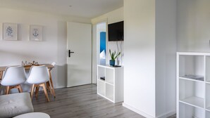 A modern, minimalist living room with a dining area, a flat-screen TV mounted on the wall, and a bookshelf.