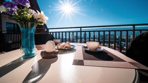 Una mesa de comedor con un jarrón de flores, platos y tazones en un balcón con vista a la ciudad y sol de fondo.