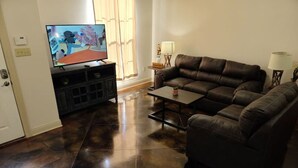 A living room with a dark brown leather sofa, a flat-screen TV on a stand, and a coffee table. - Chateau 18, spacious townhouse in Tickfaw, LA (Tickfaw)