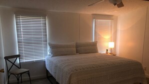 Une chambre à coucher avec un lit, deux fenêtres équipées de stores, une chaise, une table de chevet avec une lampe et un ventilateur de plafond.