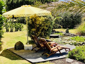 A garden with a wooden deck, two rocking chairs, a yellow umbrella, and a pond with lily pads. - Studio (Luz)