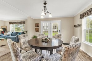 A dining area with a round table and patterned chairs, a chandelier, and a view of the outdoors through large windows.