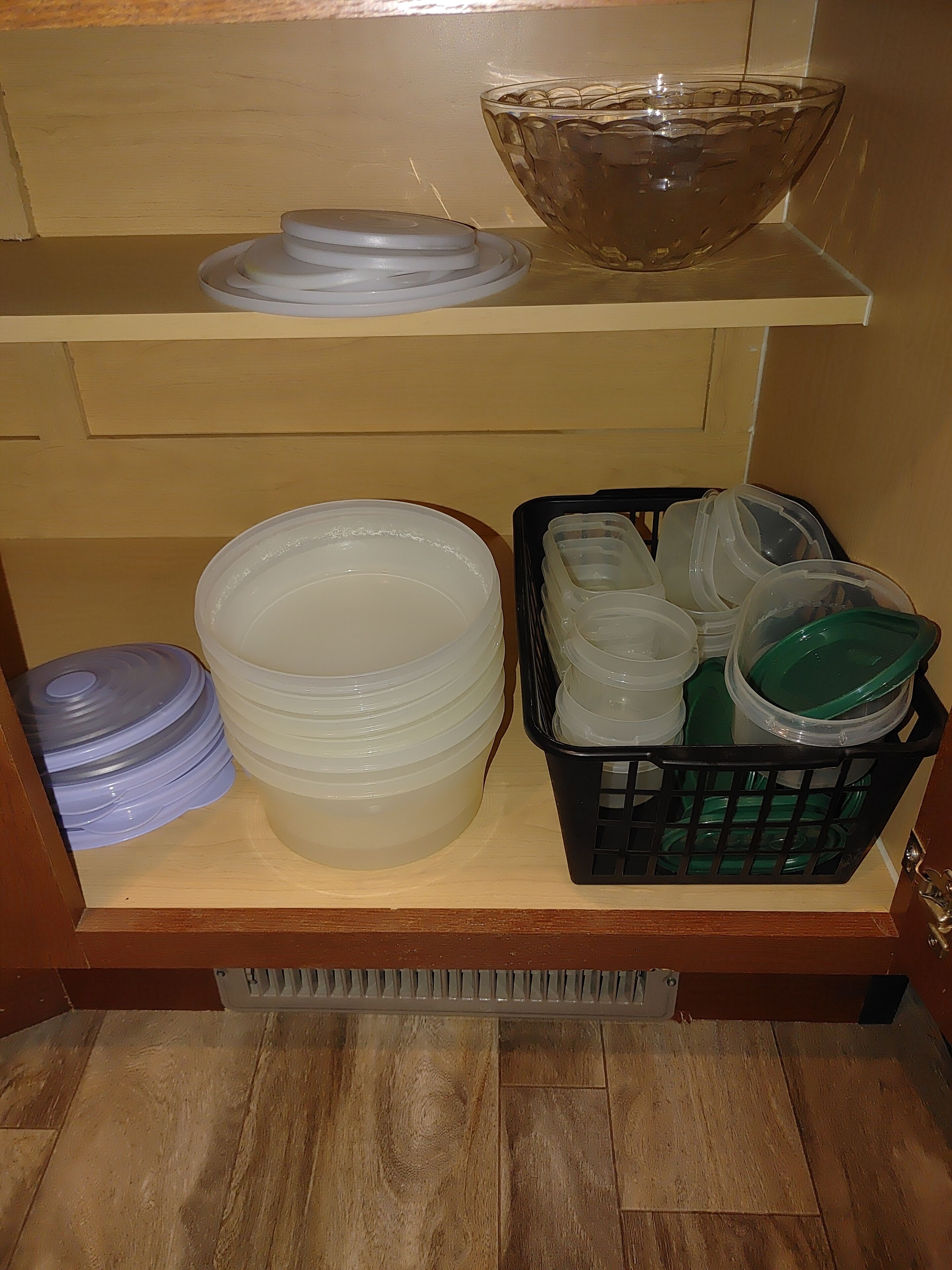 A kitchen shelf with stacked plastic containers, a glass bowl, and a black basket with green lids.