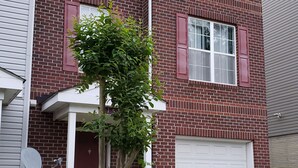 A two-story brick house with a garage, a small tree in the front yard, and two trash bins. - DC  Townhouse  close to Capitol (Washington)