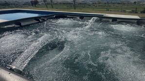 Un jacuzzi con agua en movimiento, rodeado por una piscina y una vista de una zona residencial con casas y montañas al fondo.