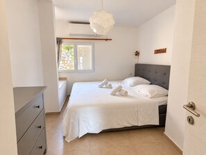 A bedroom with a bed, a dresser, a window with blinds, a wall-mounted air conditioner, and a wall-mounted light fixture.