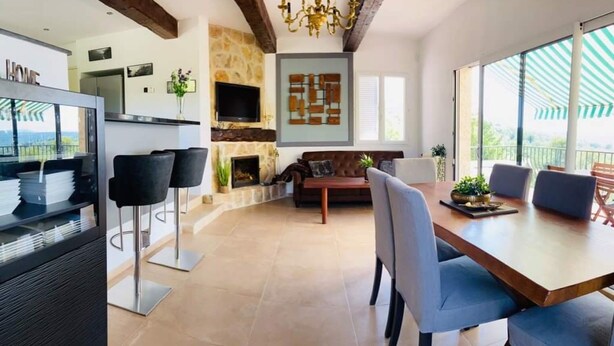 A modern living room with a fireplace, a dining area with a wooden table and chairs, and a kitchen with a stone accent wall and a TV mounted above it.