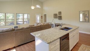 Une cuisine spacieuse dotée d’un grand îlot central, d’un four encastré et d’un évier. Le salon adjacent est agrémenté d’un canapé beige et d’un ventilateur de plafond.