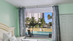 Una habitació d'hotel amb un llit, vistes a una piscina i palmeres, i una porta.