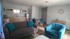 Un saló modern amb un sofà, una butaca blava, una taula de menjador amb cadires, una zona de cuina amb armaris i electrodomèstics, i un rellotge a la paret.