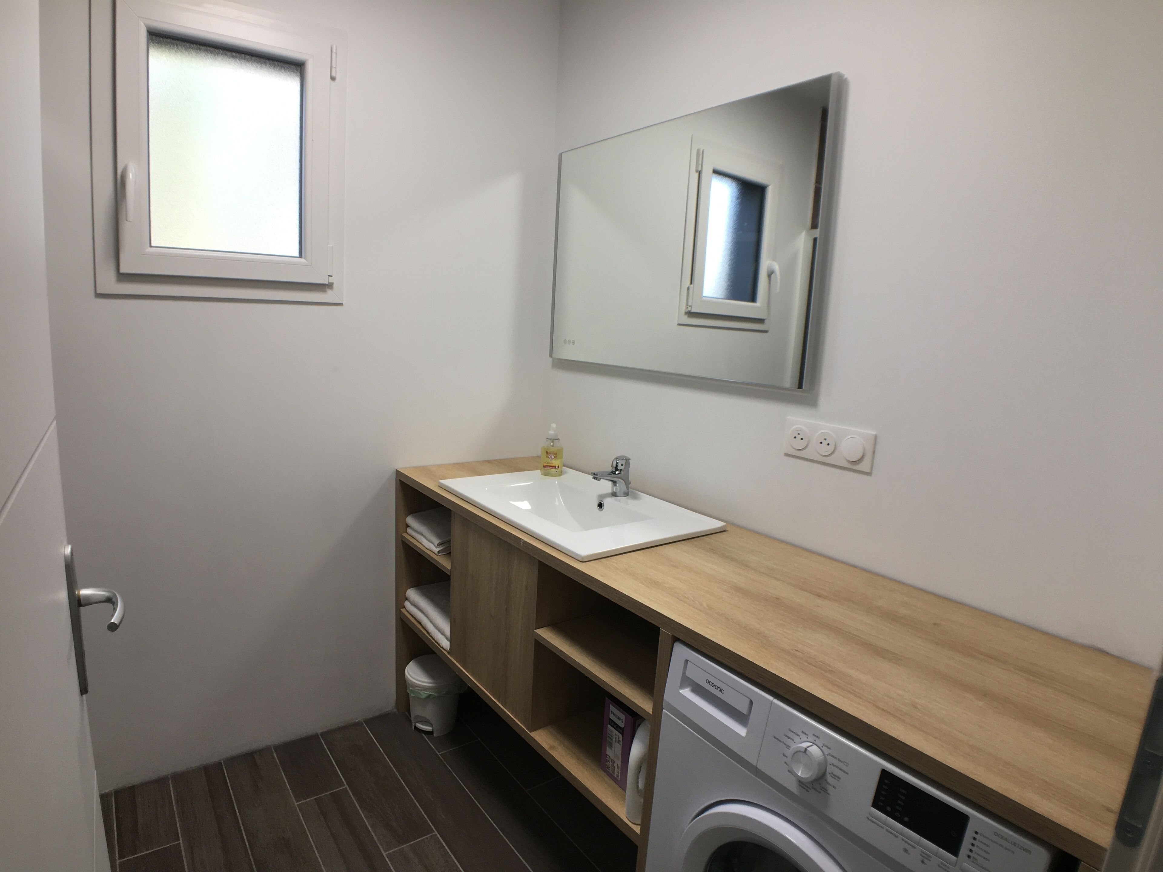 A compact bathroom with a white sink, wooden vanity, a mirror, a washing machine, and a small window.