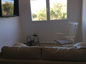 Une pièce avec un canapé beige, une chaise blanche, une petite table avec une télécommande et un téléviseur fixé au mur.