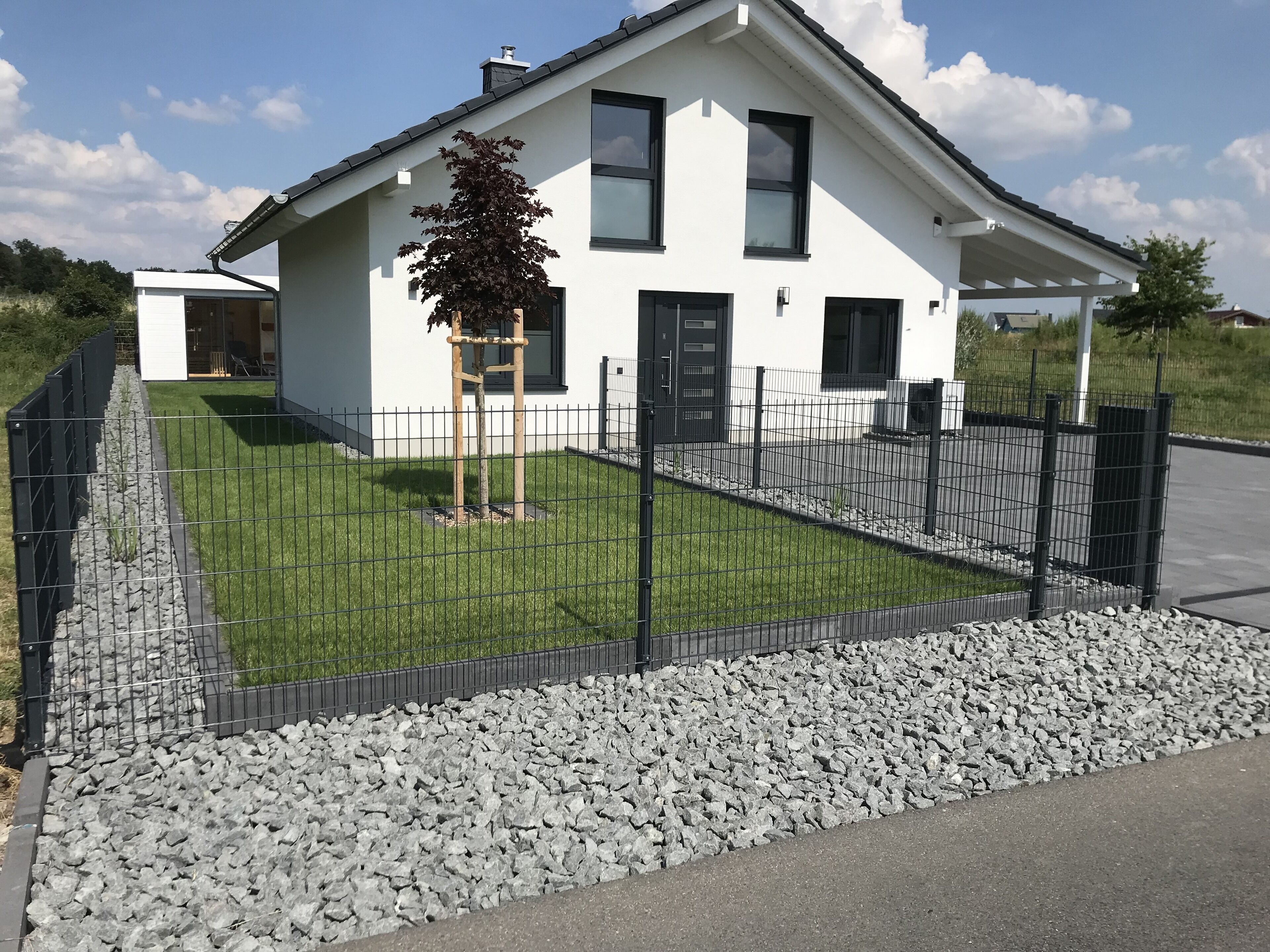 Une maison moderne à deux étages, avec une façade blanche, une clôture noire et un petit arbre dans le jardin.