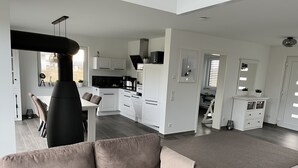Een moderne woonkamer met een bank, een eethoek met een tafel en stoelen, een keuken met witte kasten en zwarte apparaten, en een gang die naar andere kamers leidt.