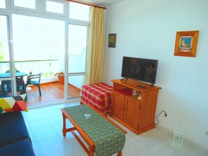 Ein Zimmer mit Fernseher auf einem Holzboden, einer Sitzbank, einem Tisch mit Stühlen und einem Balkon mit Blick.