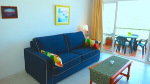 Una habitación con un sofá azul, una mesita, un cuadro en la pared y una ventana con cortinas.