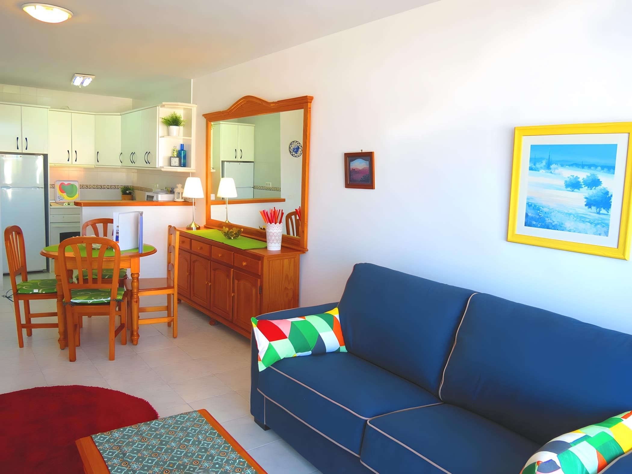 Una sala de estar con un sofá azul, un comedor con mesa y sillas, y una cocina con armarios y un refrigerador.