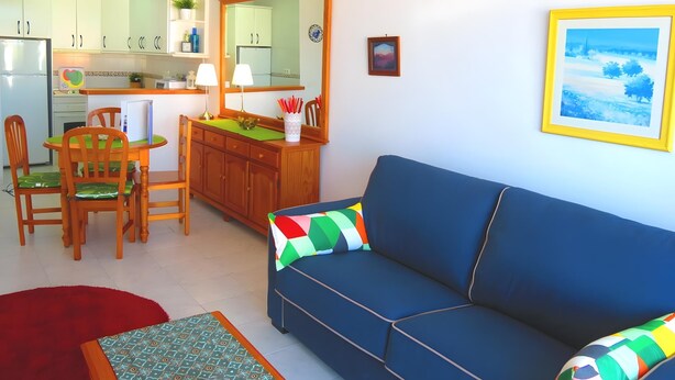 Una sala de estar con un sofá azul, un comedor con mesa y sillas, y una cocina con armarios y un refrigerador.