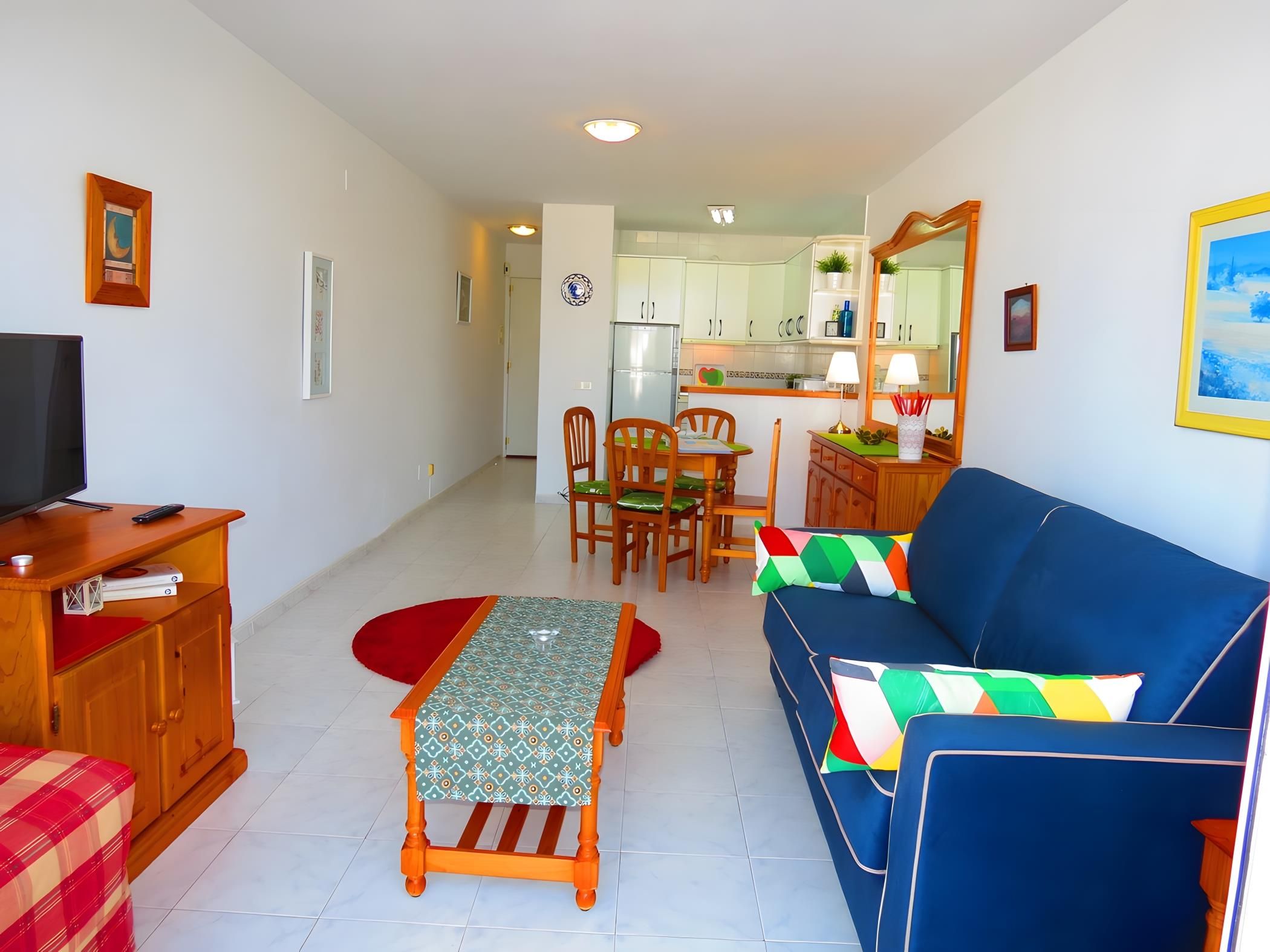 A living room with a sofa, a TV, a dining table with chairs, and a kitchen in the background.