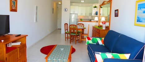 A living room with a sofa, a TV, a dining table with chairs, and a kitchen in the background.