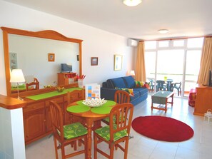 Ein modernes Wohnzimmer mit einem blauen Sofa, einem Esstisch mit Stühlen, einer Küchenzeile inklusive Fernseher und einem großen Fenster mit Vorhängen.
